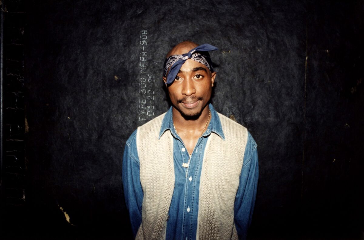 Tupac, wearing a blue and white shirt with blue bandana, stands against a black wall.