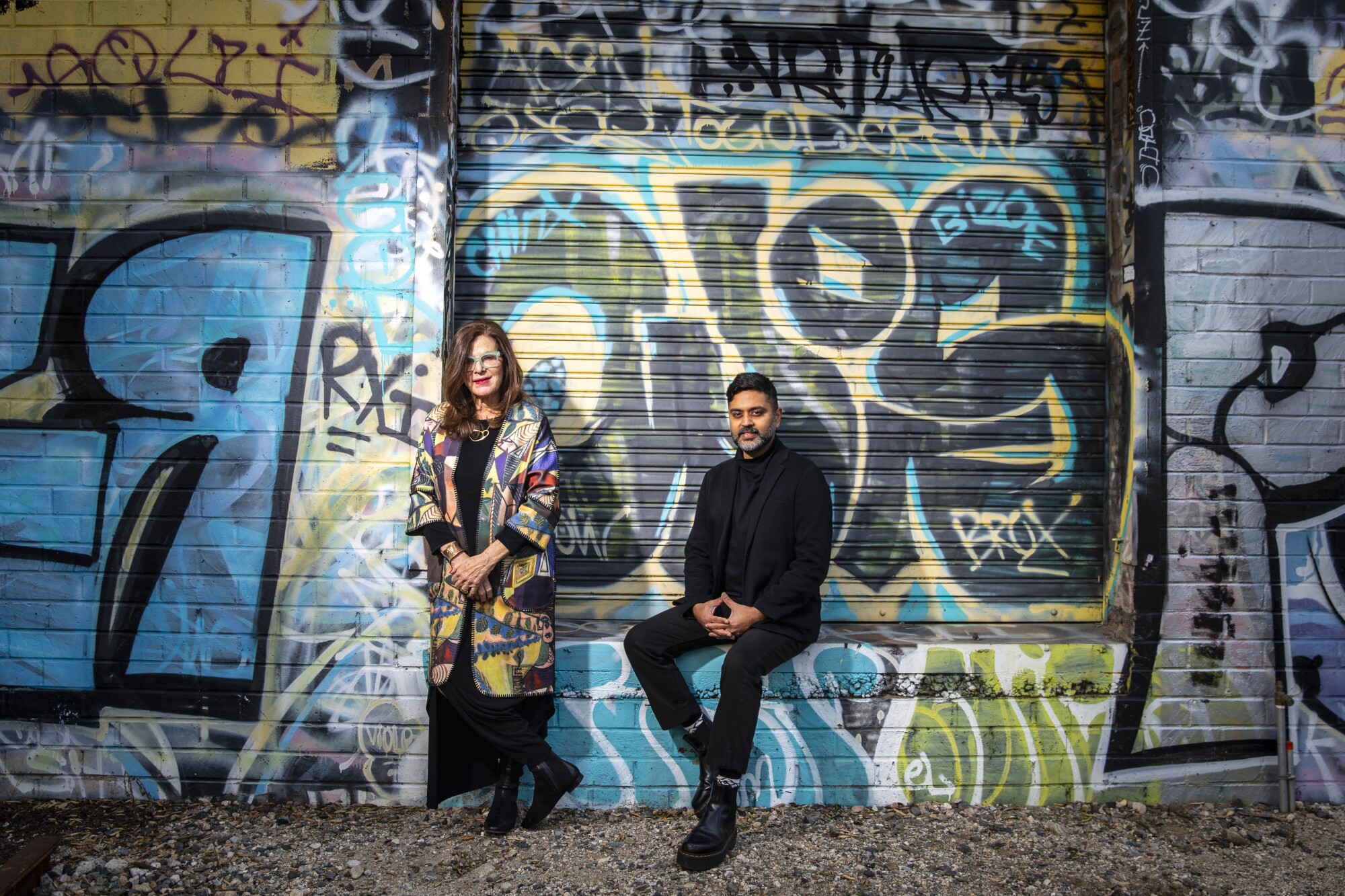 Mia Lehrer, in a bright coat, and Kush Parekh, dressed in black, stand before a graffiti covered wall