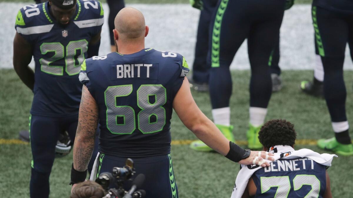 Seattle center Justin Britt (68) stands with his hand on defensive end Michael Bennett, who is seated druing the national anthem on Friday.