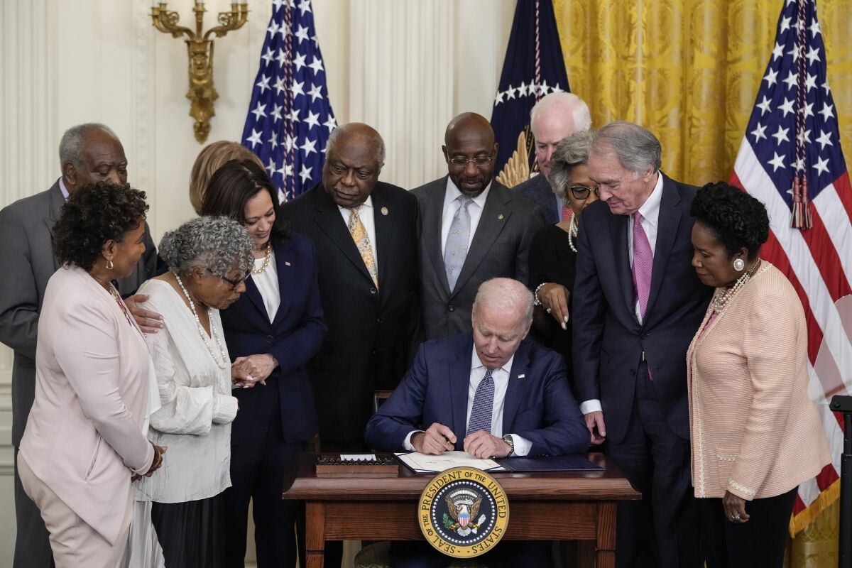 President Biden signs a document at a small desk surrounded by supporters