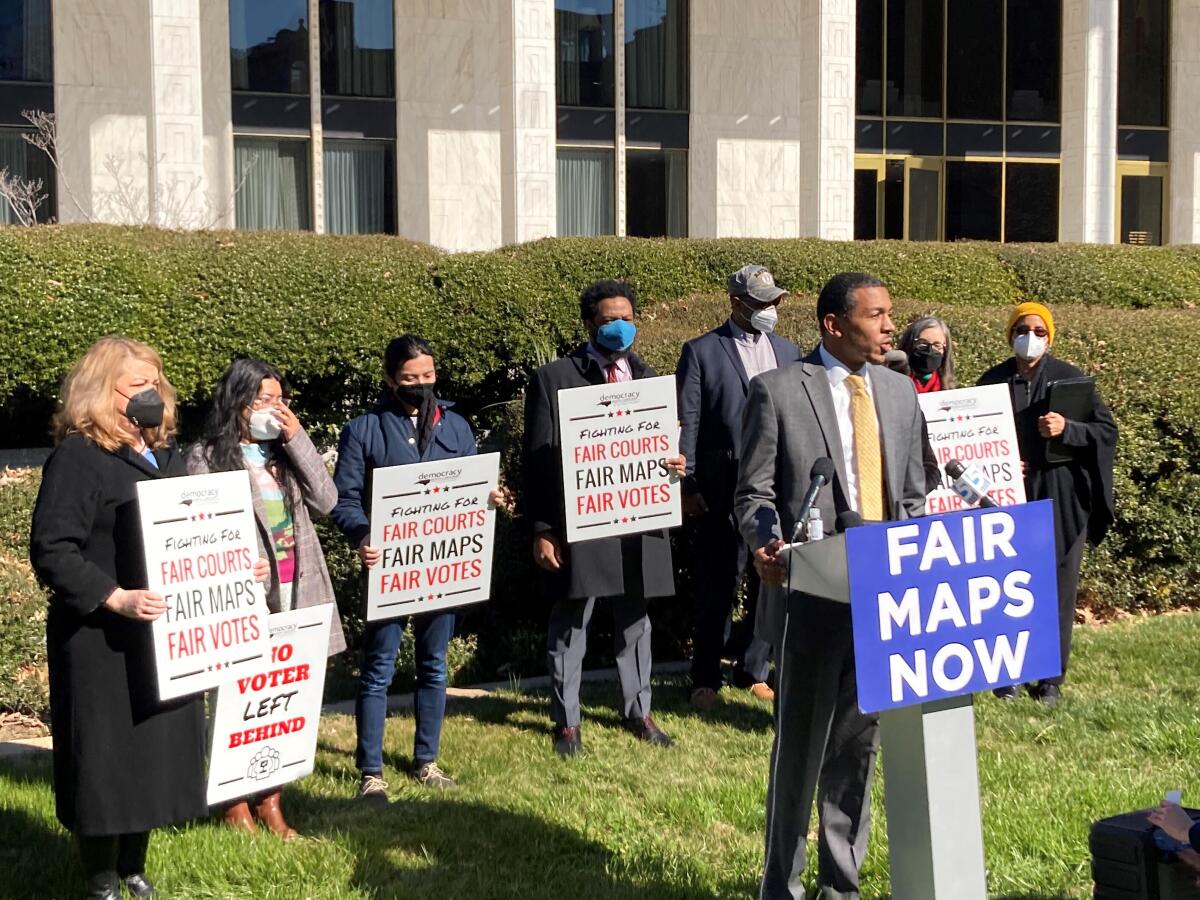 A man speaking outside a building from a lectern with a "Fair maps now" sign as several people behind him hold similar signs.