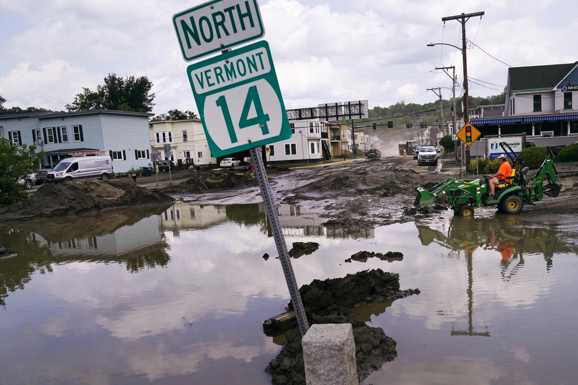A small tractor travels through mud next to floodwaters that partially submerge a sign reading "North Vermont 14."