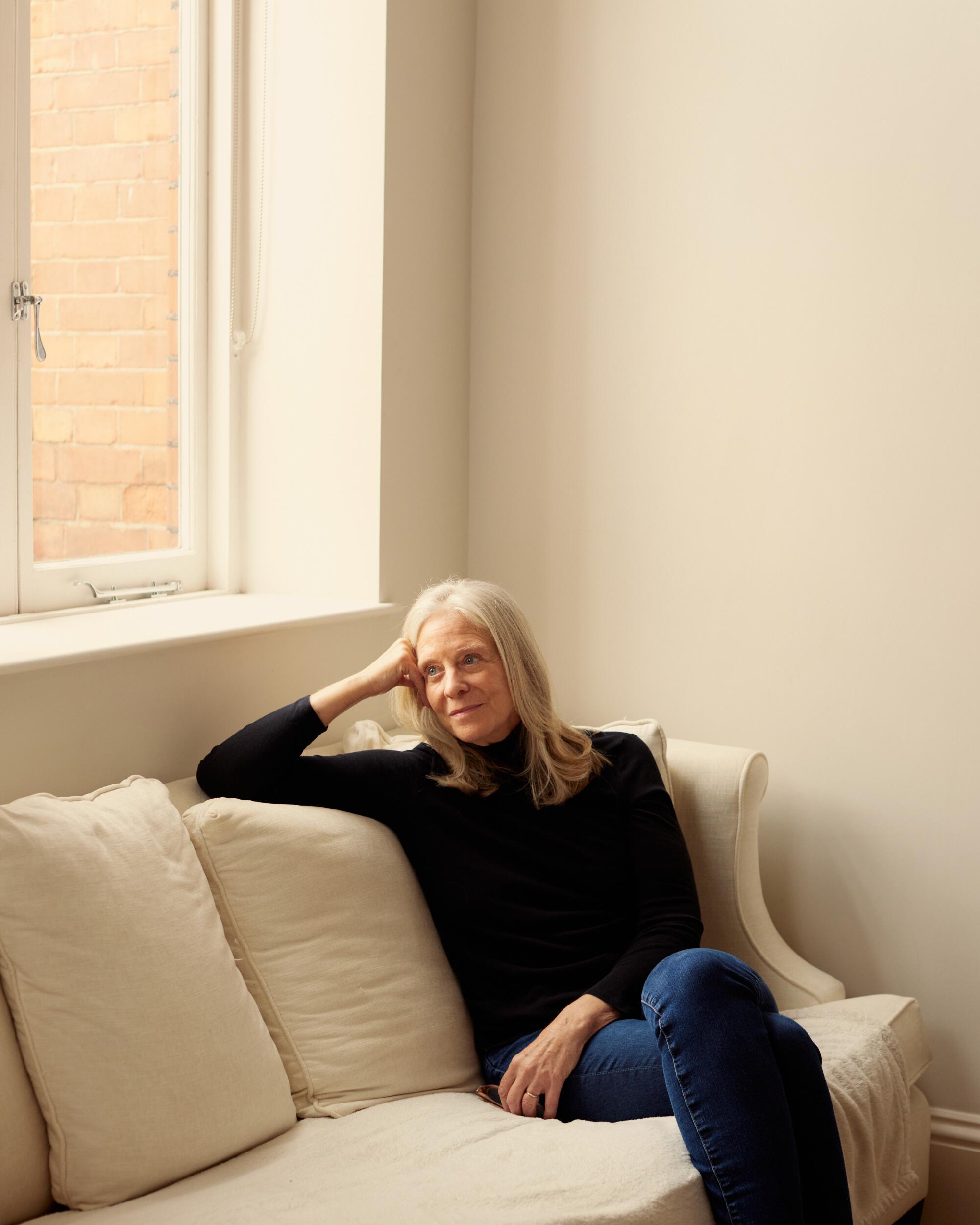 Bonnie Garmus sits on a white couch near a window in a black turtleneck and jeans.