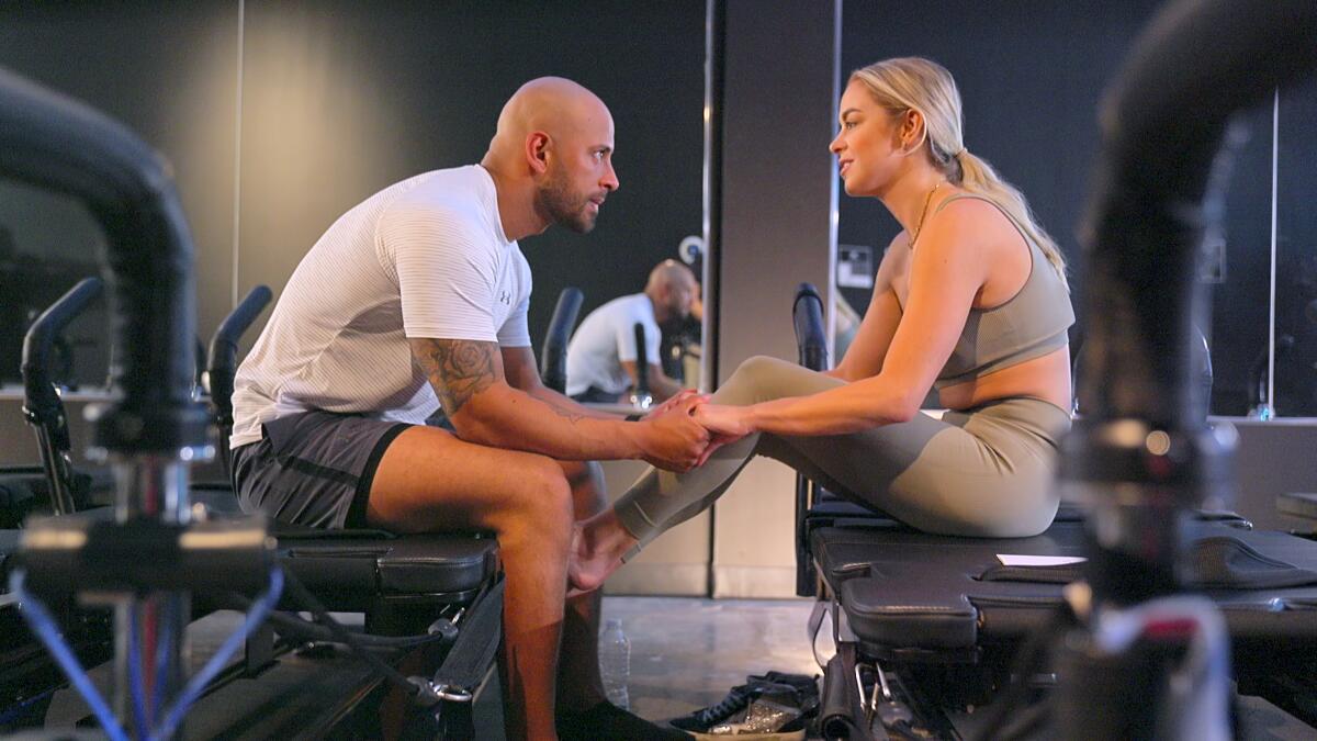 In a gym, a man in a white T-shirt and shorts holds the hands of a blond woman in a green sports bra and tights.
