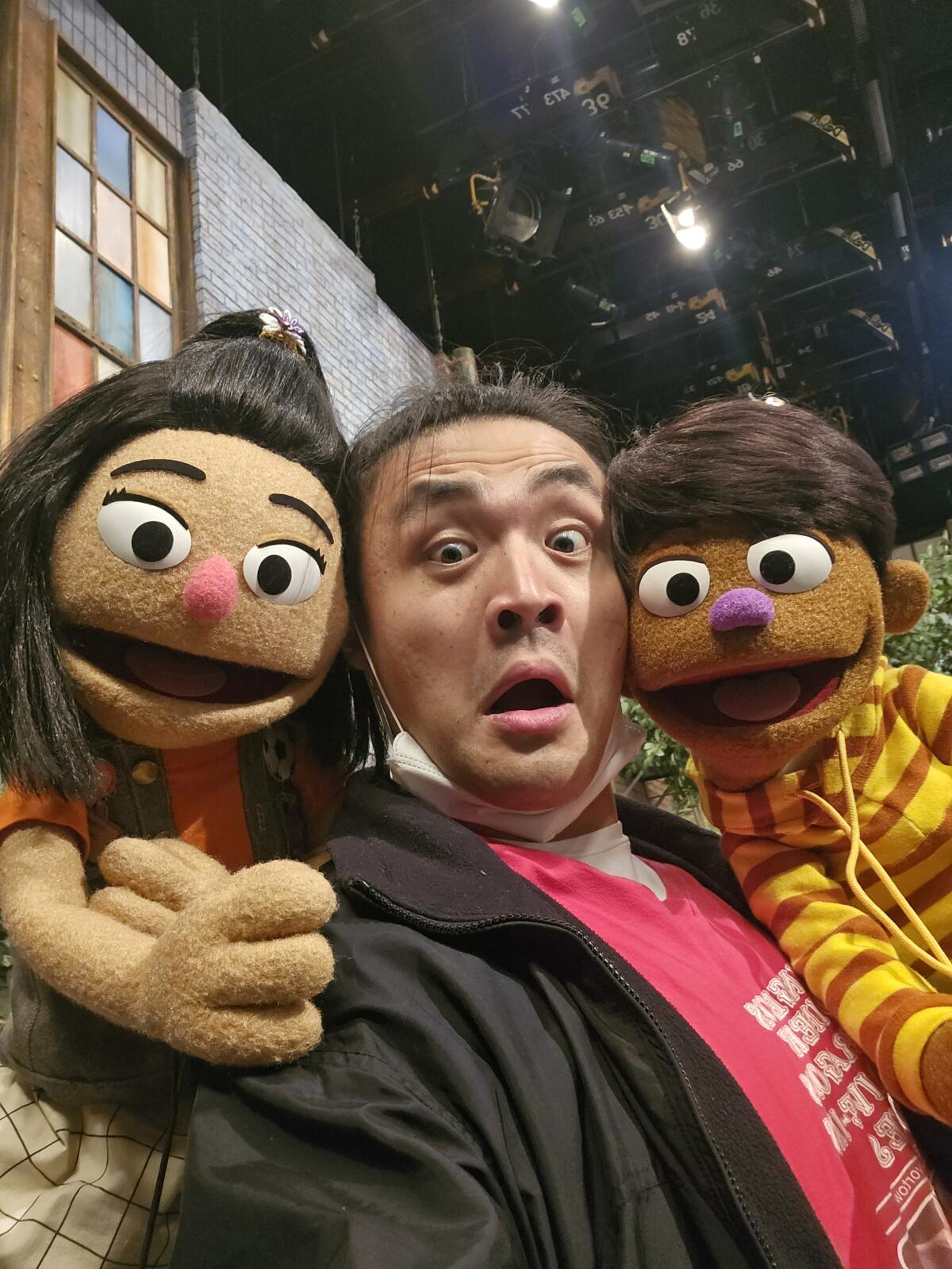 A man poses with two Muppets on set.