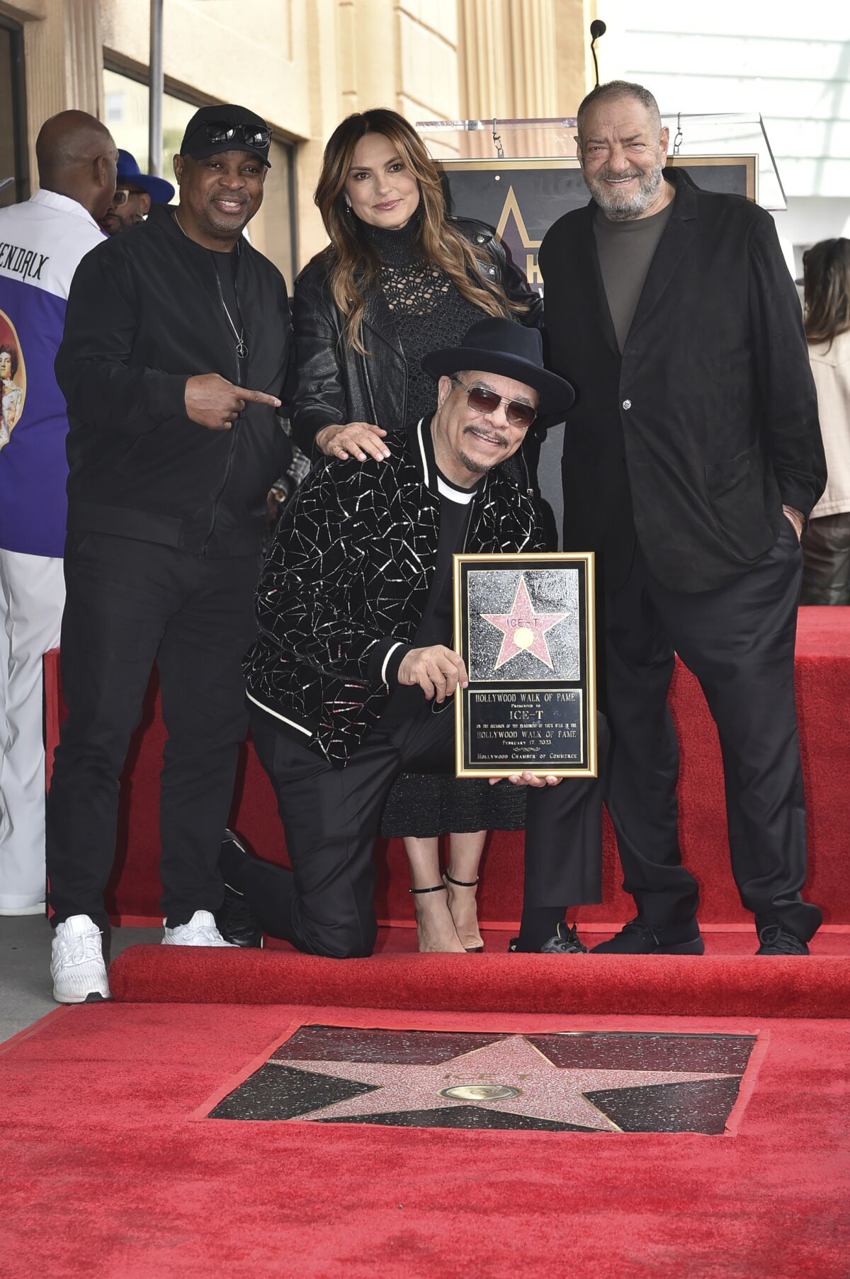 Two men with a woman between them stand behind a man in sunglasses and a hat who is getting a star on Hollywood Boulevard