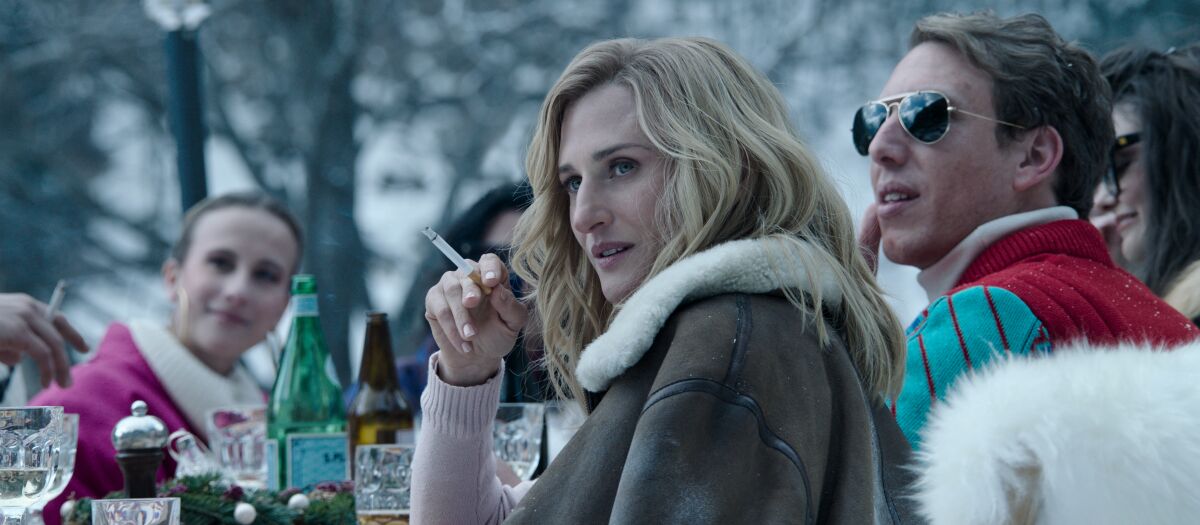 A blond woman smoking and sitting at a table with others, outside in the snow.