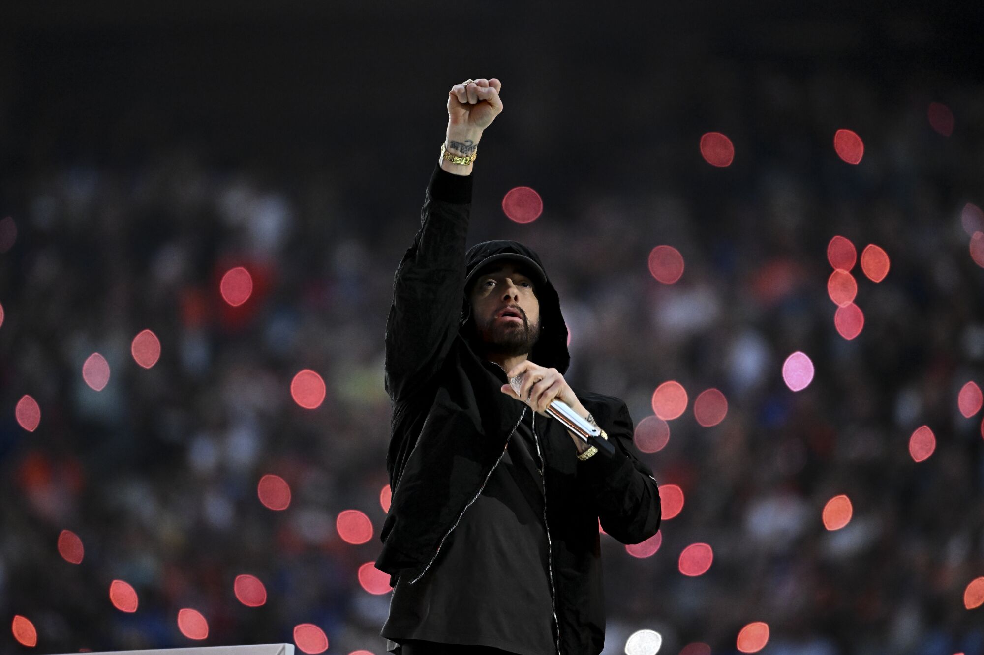 A man in a black hoodie holds a microphone in one hand and holds the other up in a salute.