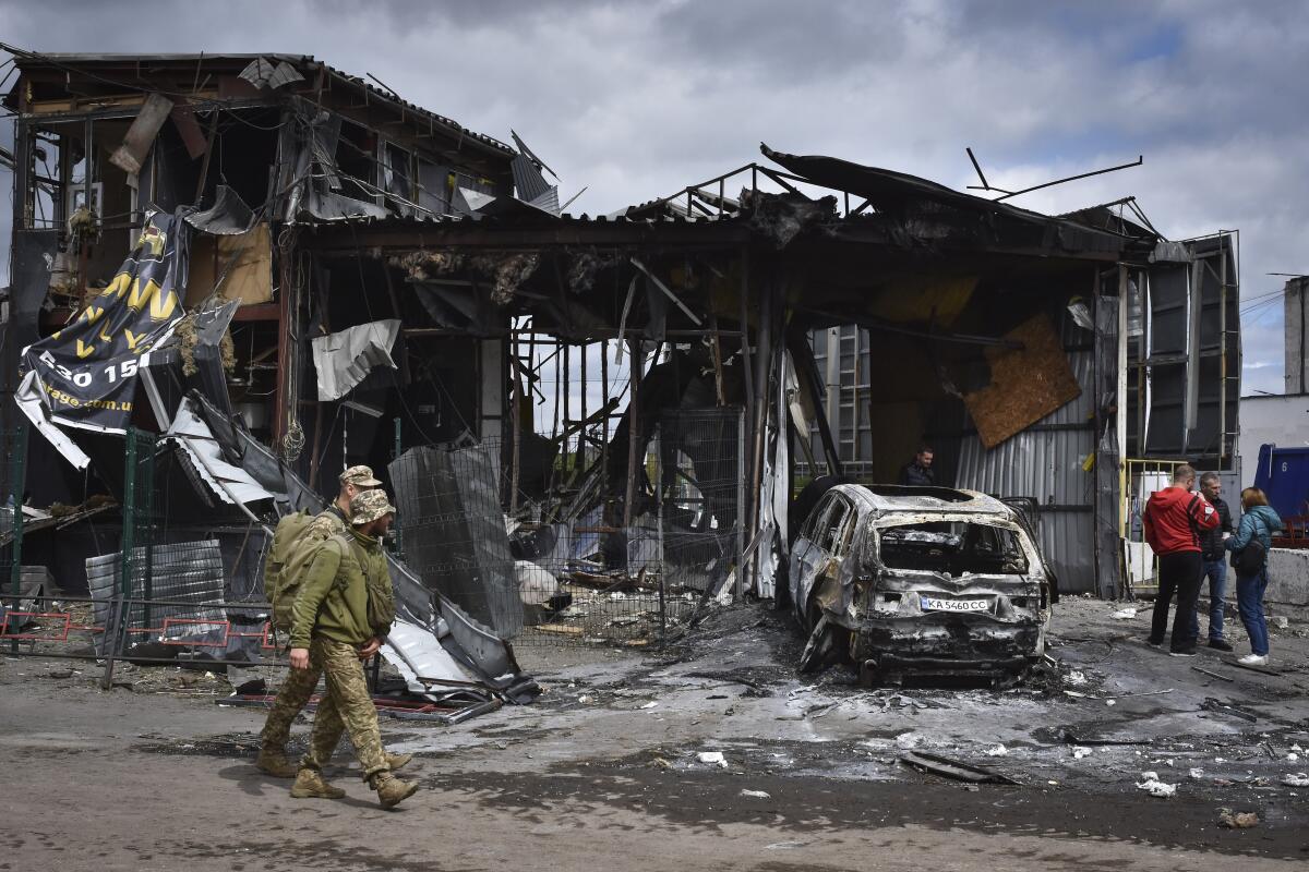 Two soldiers in camouflage walk by a charred, gutted building and car where three people in civilian clothes huddle and talk