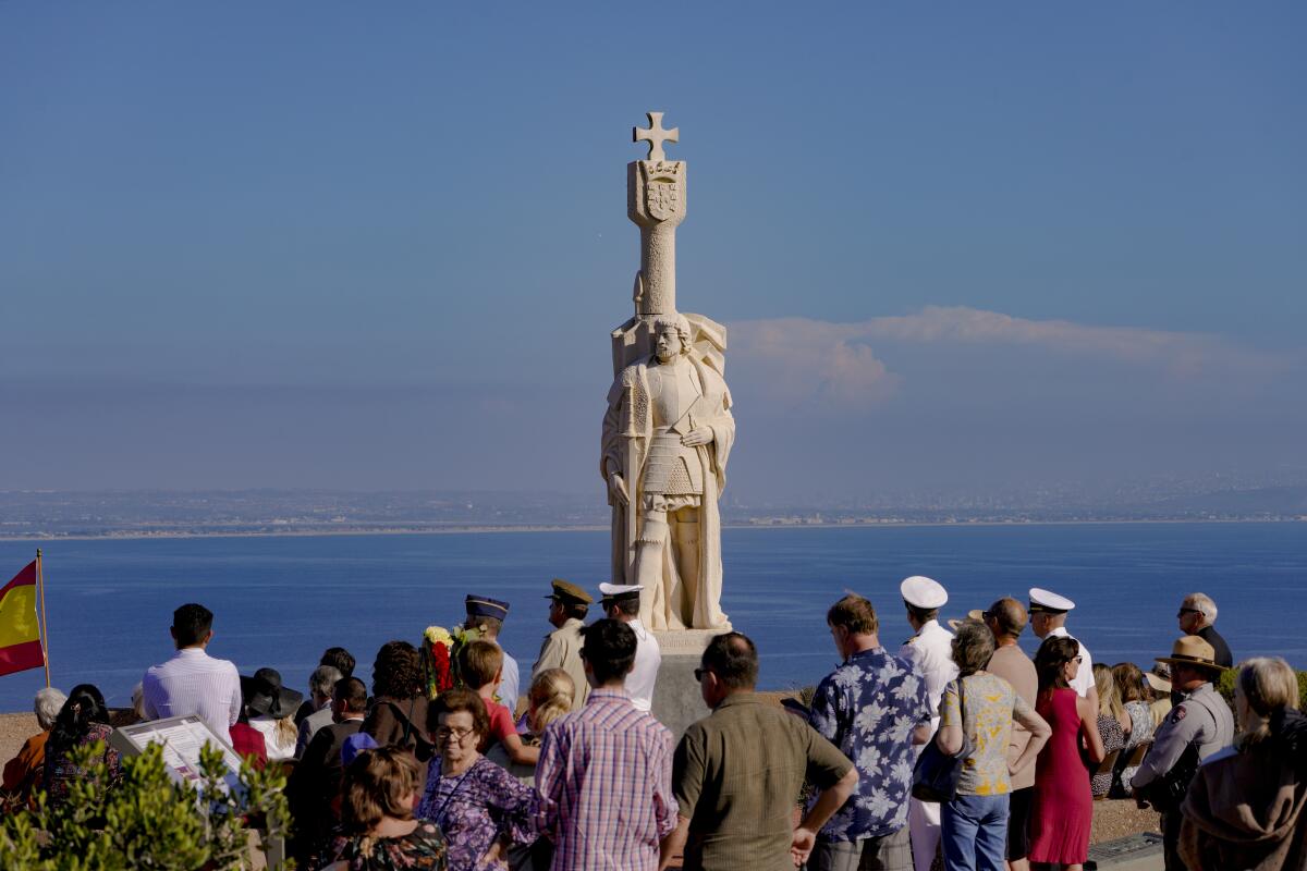 A crowd of people, some in naval uniform, surround a monument overlooking the ocean.