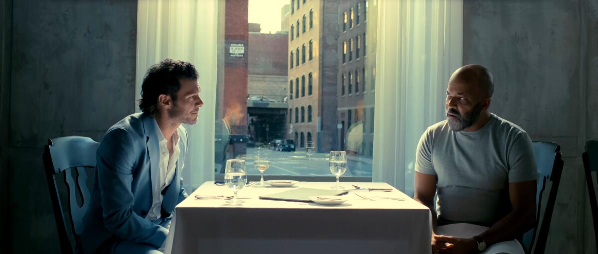Two men seated in a restaurant next to a window. One looks warily across the table at the other.