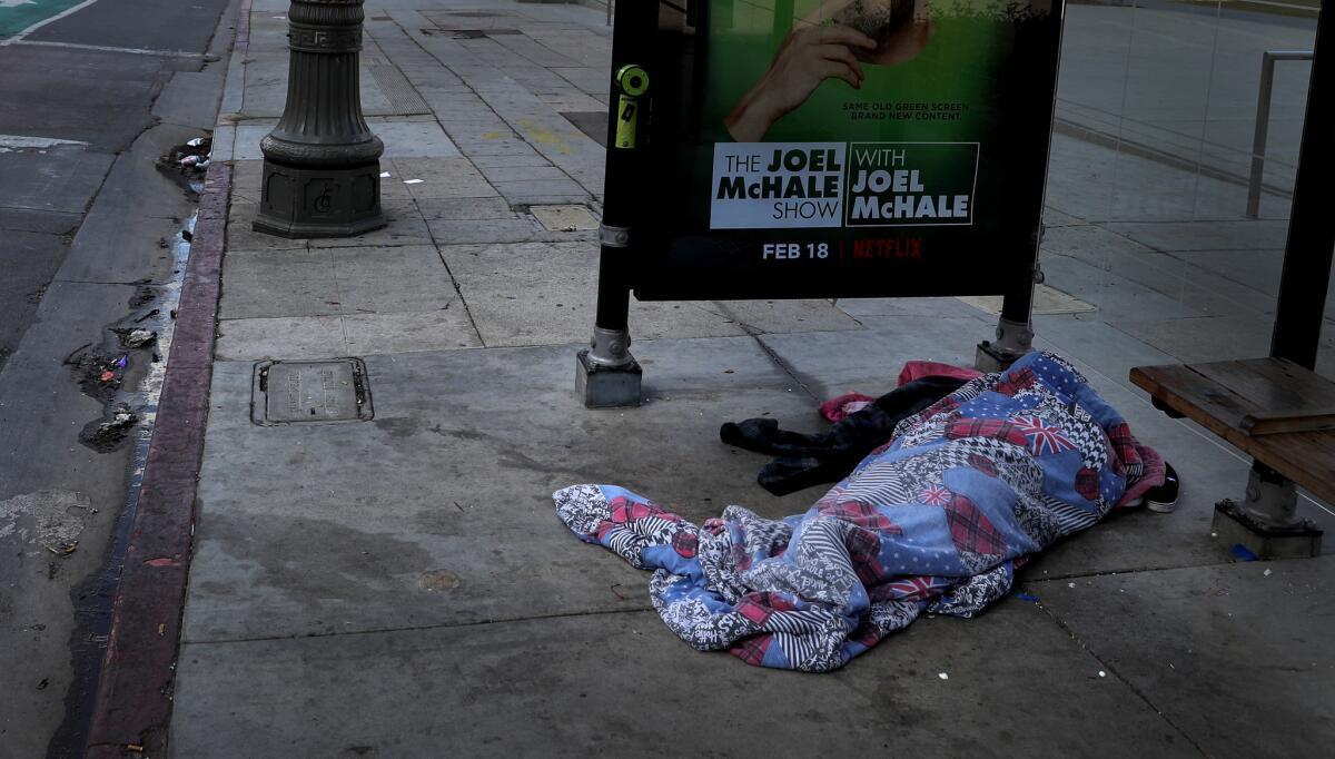 A homeless person wrapped in a blanket sleeps on the street under a bus stop across from Los Angeles City Hall.