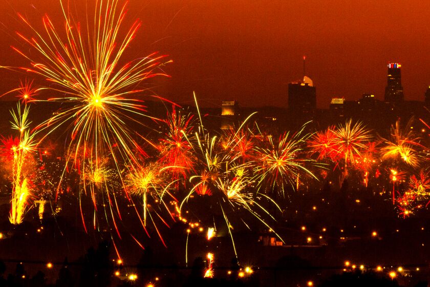 Fireworks exploding into an orange sky. A dark tinted Los Angeles skyline can be seen behind the fireworks.