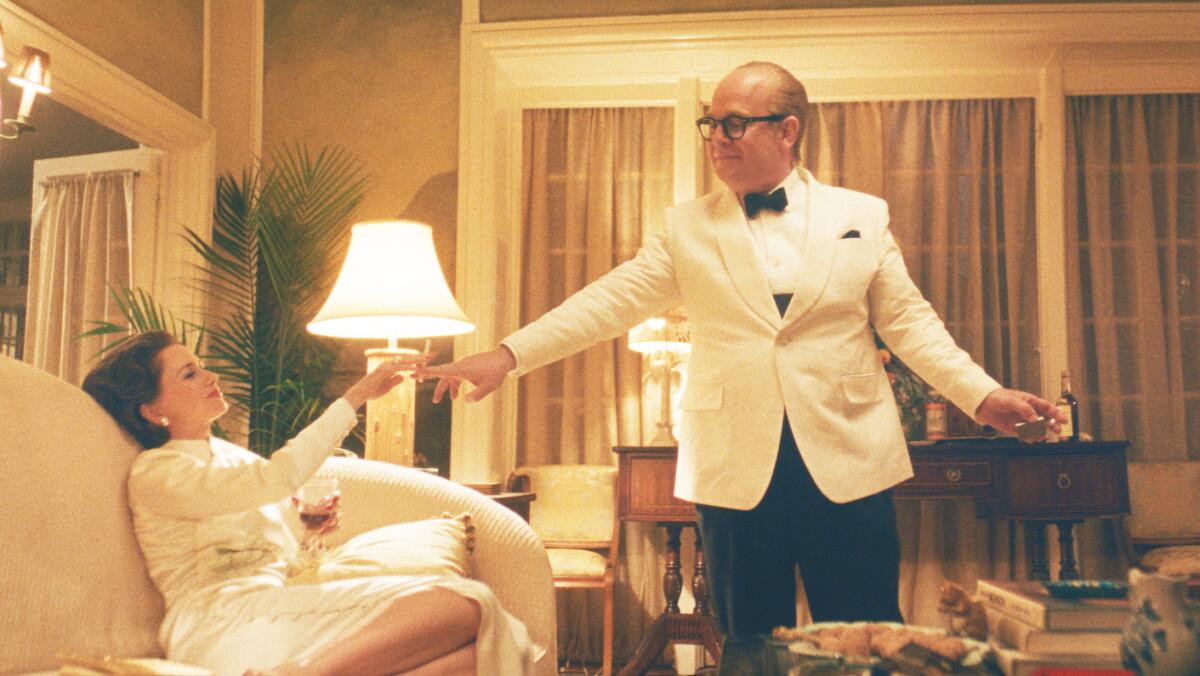 A man in a tux with white jacket stands reaching his hand toward a woman dressed in white lounging on a white sofa