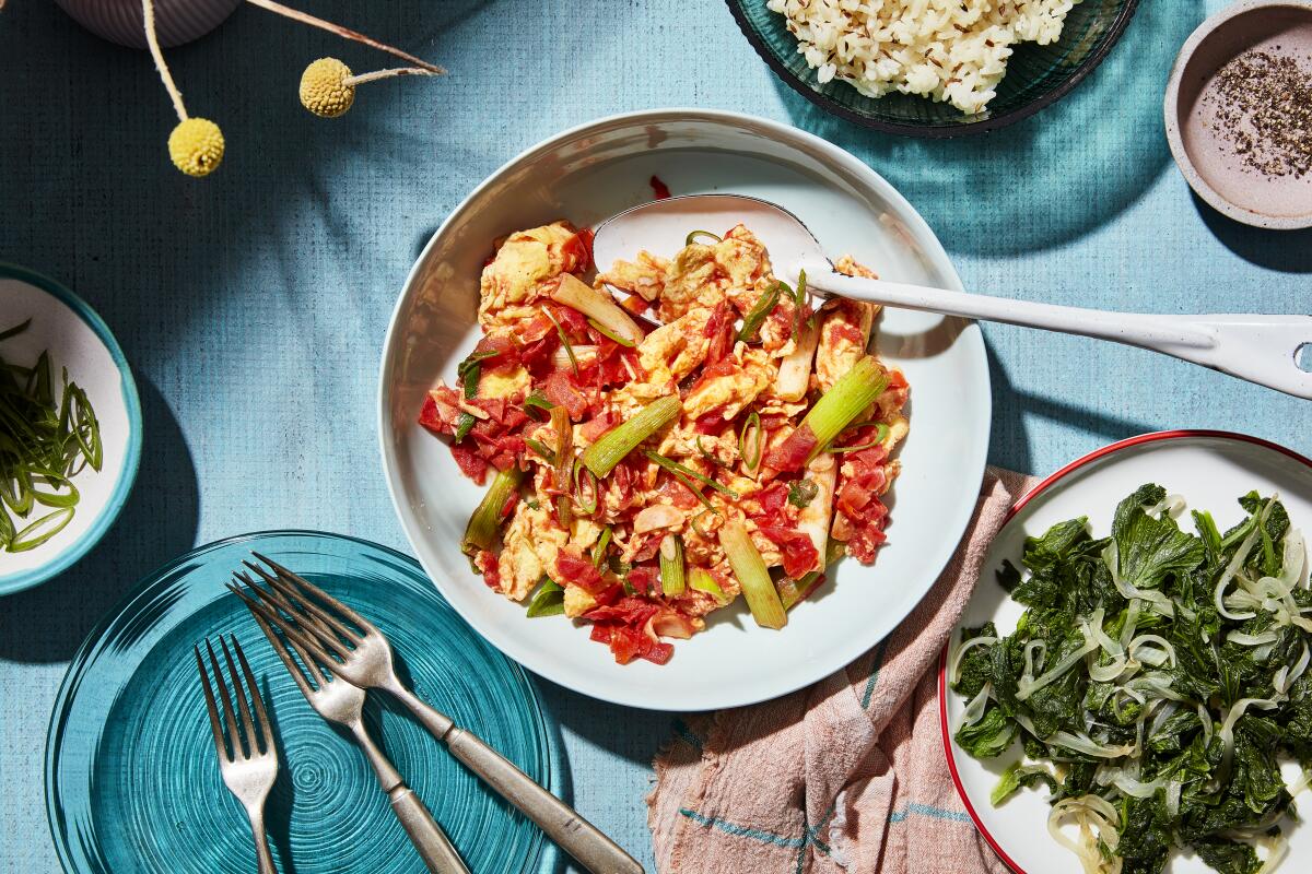 Overhead view of a plate with a mix of tomatoes and eggs and chopped green onion, alongside a plate of steamed greens.