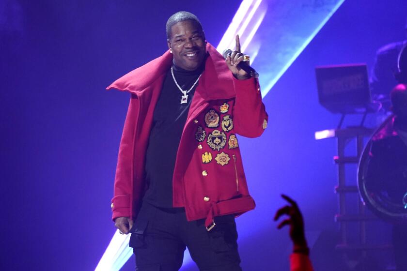 A man in a red jacket and silver necklace standing on a stage, smiling and pointing a finger upward