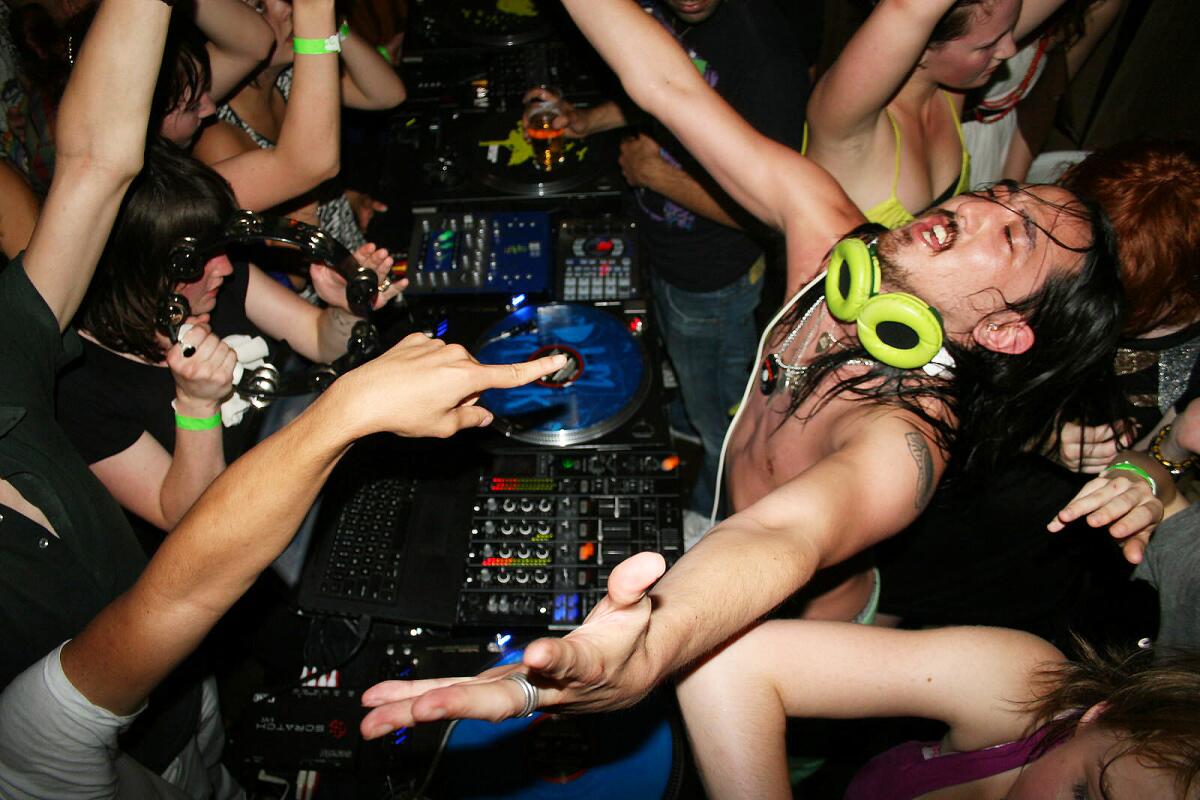 A shirtless DJ plays music at a club, surrounded by partygoers.