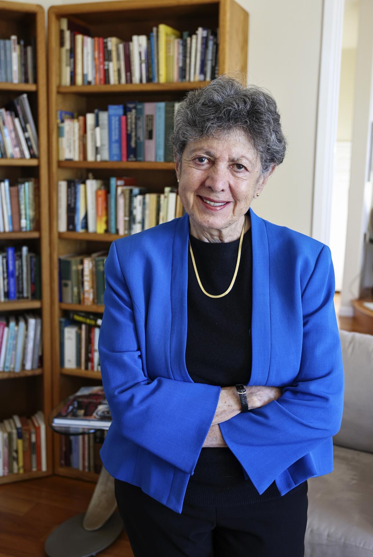 An older woman poses in a blue jacket, arms crossed, in front of a bookcase