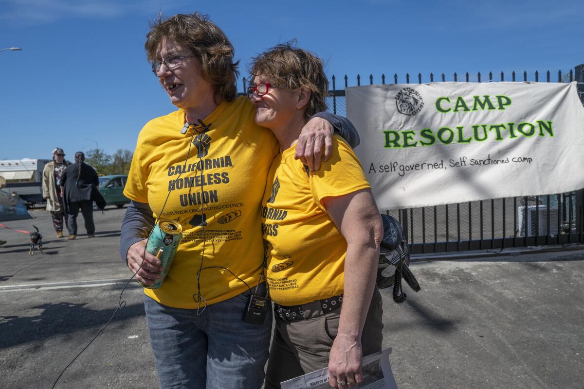 Two women smile and embrace in front of a sign that reads: "Camp Resolution, a self-governed, self-sanctioned camp."