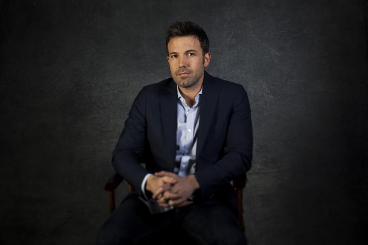 Ben Affleck poses for a portrait, seated with his hands clasped in front of a dark gray background