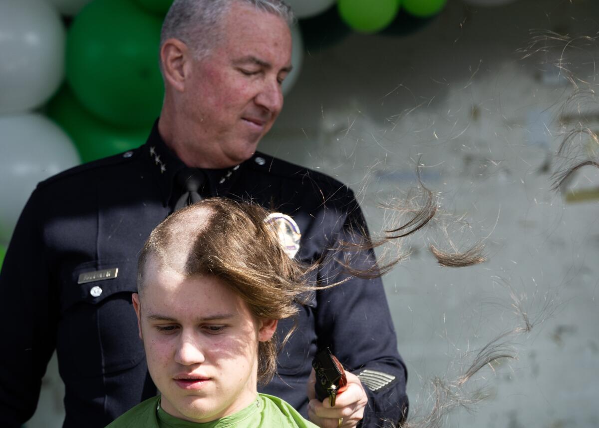 A police officer stands behind a seated youth with a half-shaved head as pieces of his hair float away on the wind.