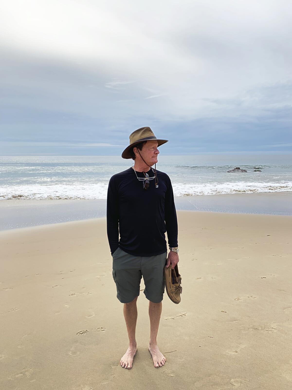 A man in shorts holds his shoes on the beach