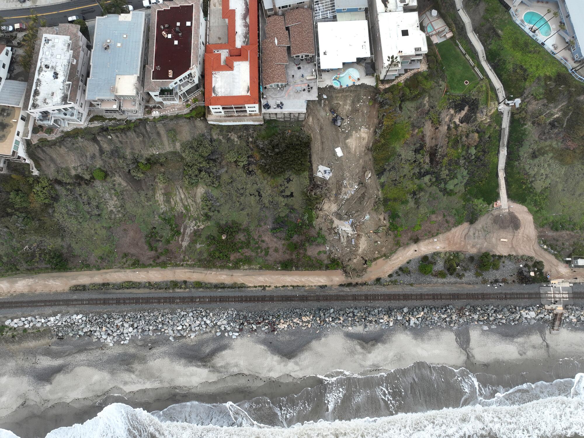 An overhead aerial view of homes on a cliff, a section where the land has given way,  and ocean.
