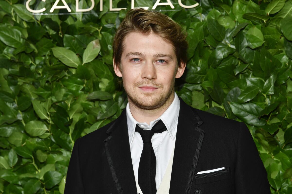 Joe Alwyn standing in front of a leafy green background in a black jacket, white button-down shirt and black tie