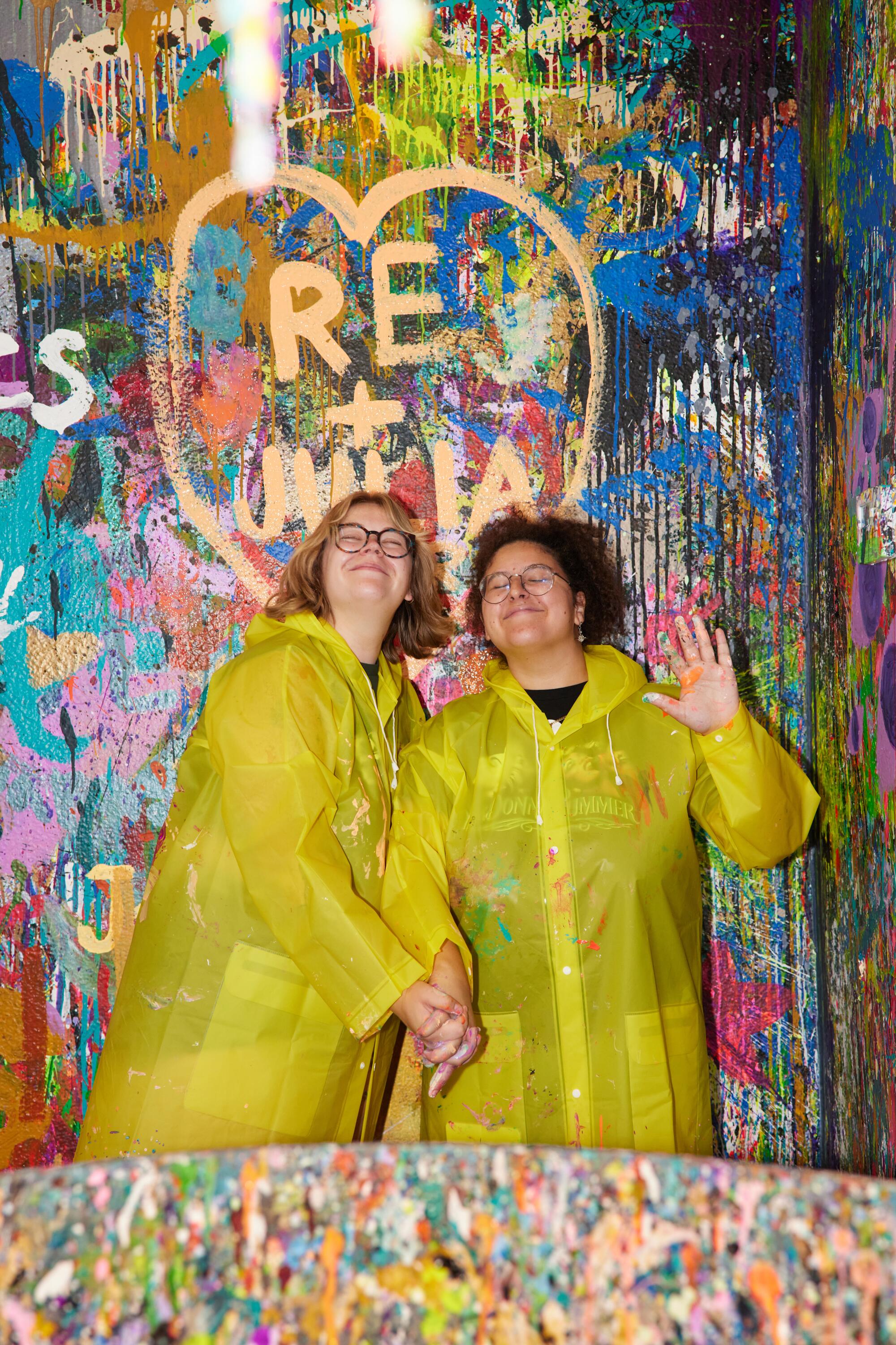 Two people wearing yellow raincoats pose in front of a heart painted onto the multicolor wall.