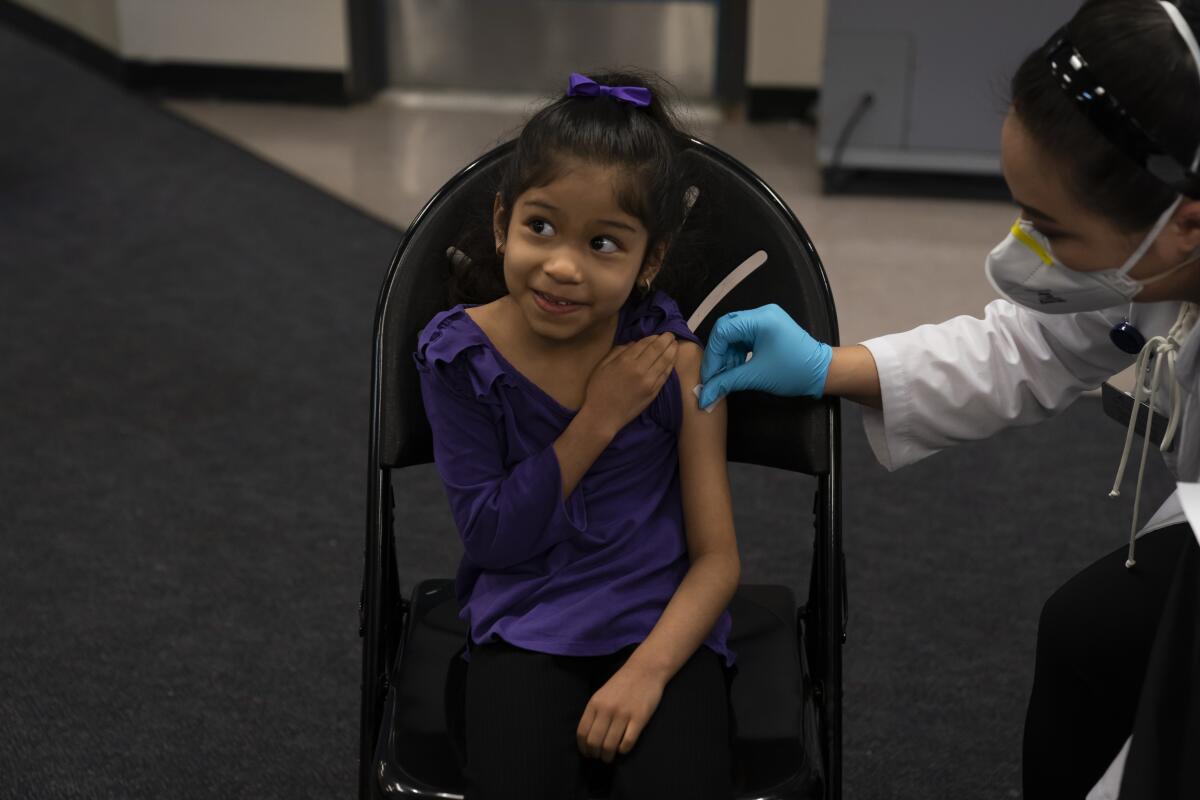 A 6-year-old girl smiles at her mother (off-camera) as a pharmacist prepares to give her a COVID vaccination