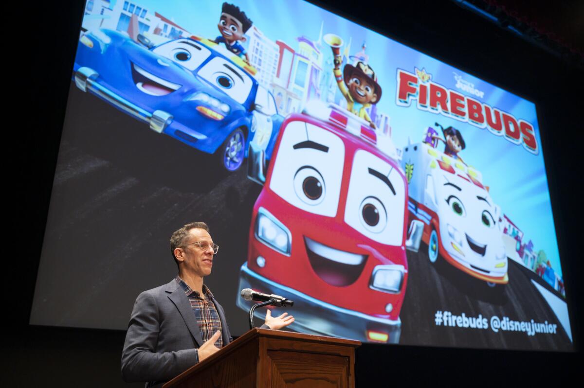 A man speaks into a microphone at a podium. Behind him is a screen showing a cartoon.