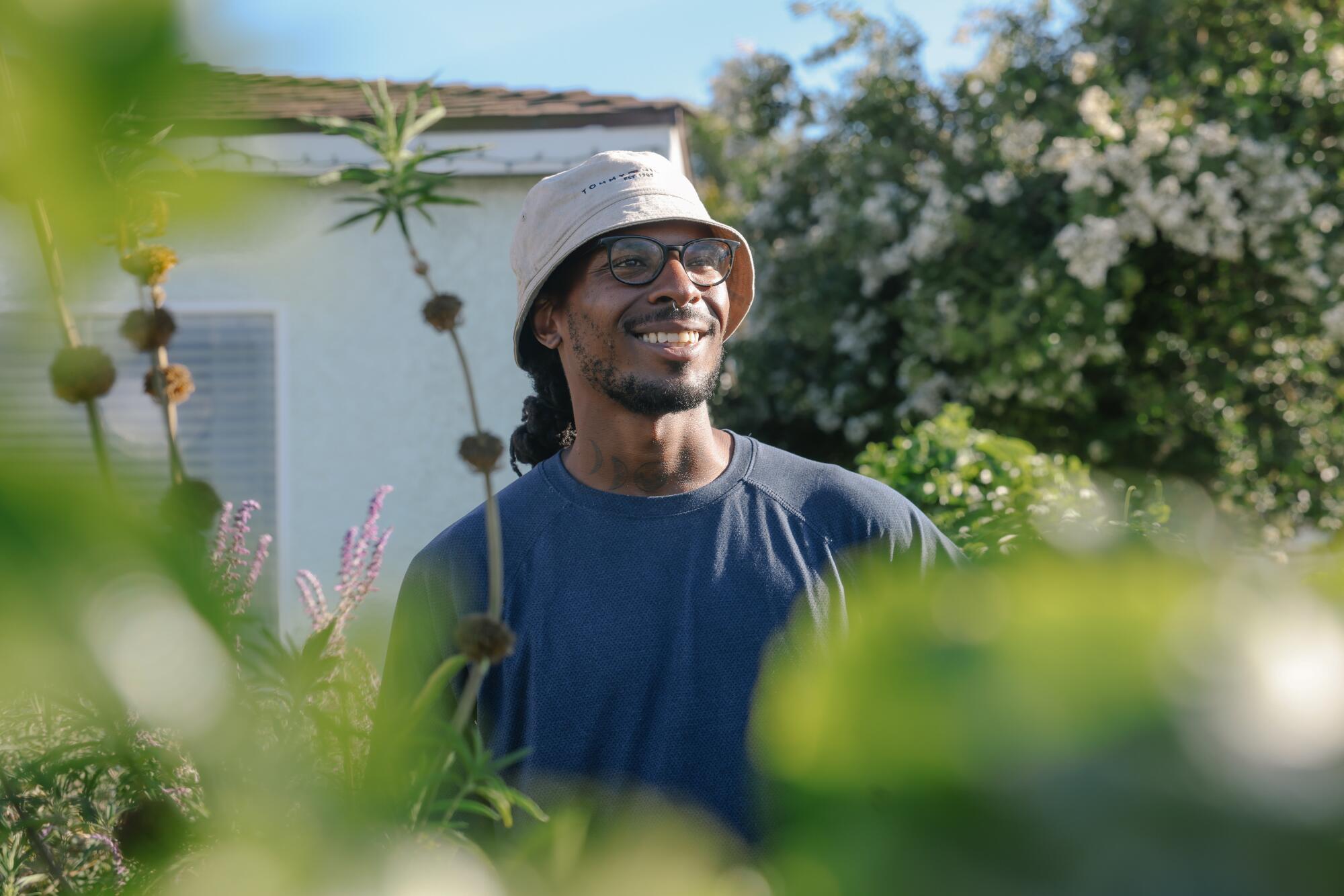 A smiling man in a bucket hat stands outside smiling, surrounded by plants