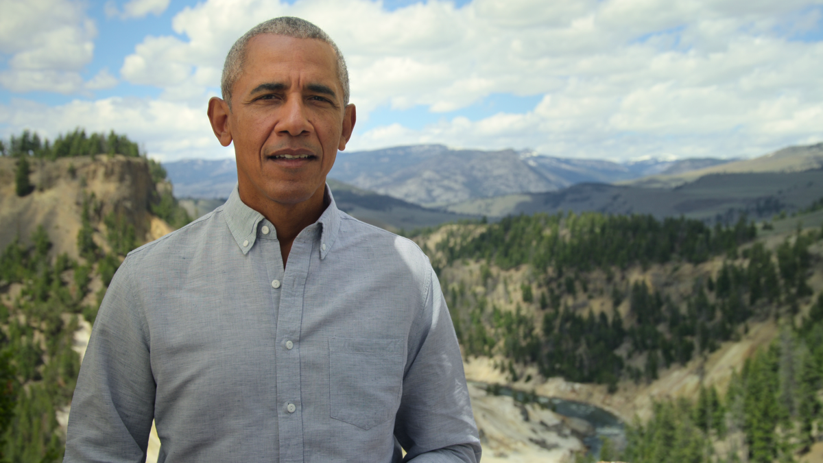 Barack Obama in a light button-down shirt stands before a sweeping vista