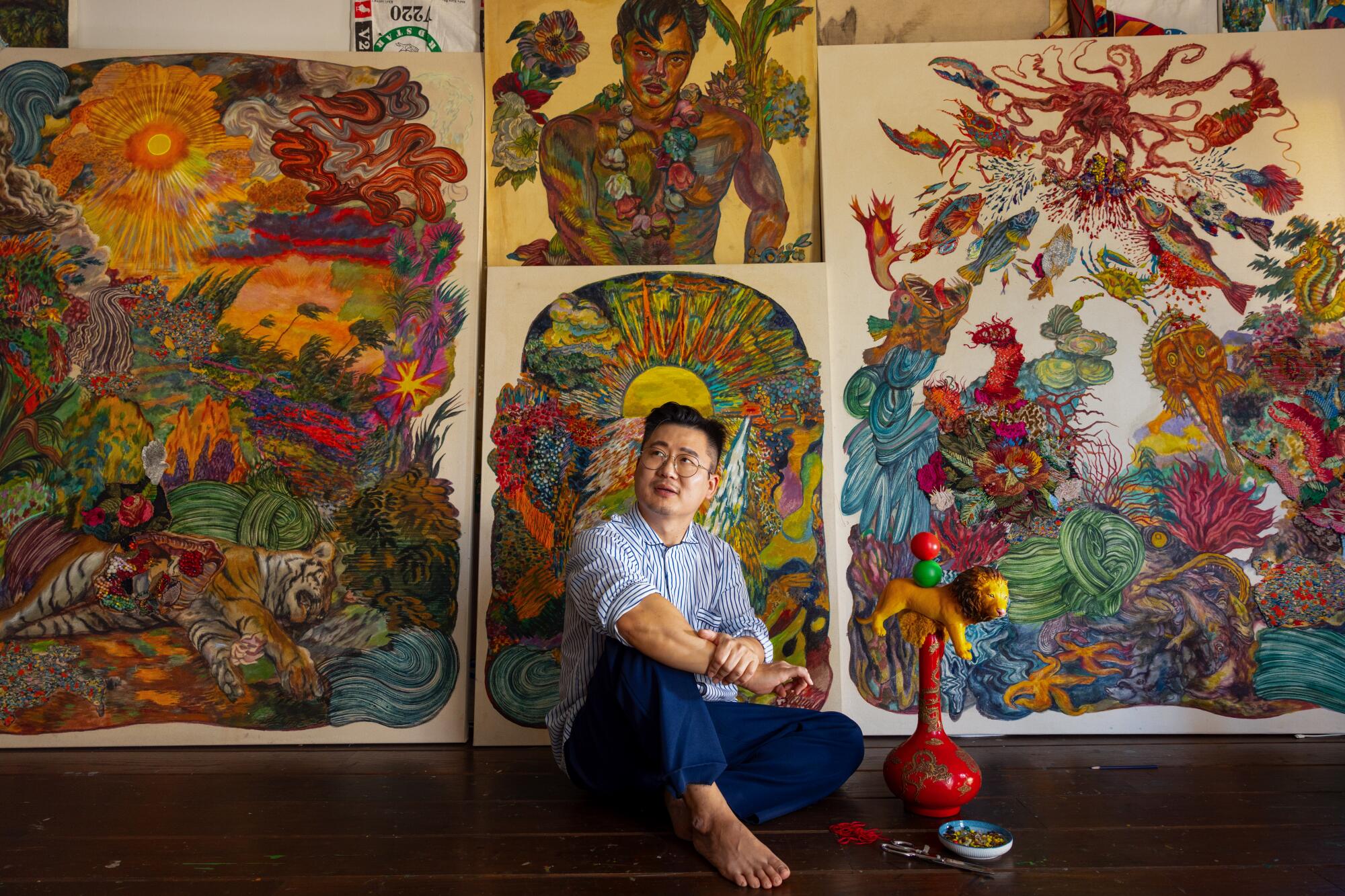 Artist Ken Gun Min sits in front of colorful paintings in his studio