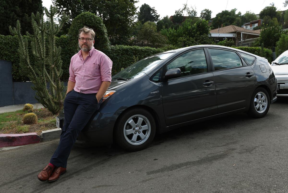 Steven Simon, posing in a pink shirt, leans against the hood of a gray Prius.