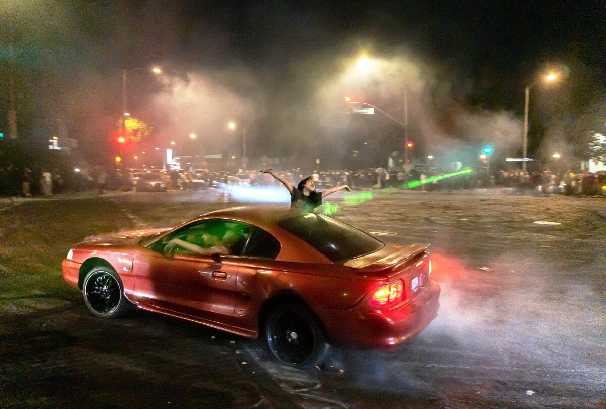 A person with arms raised hangs out of a car's passenger window as the vehicle spins in an intersection, smoke trailing.
