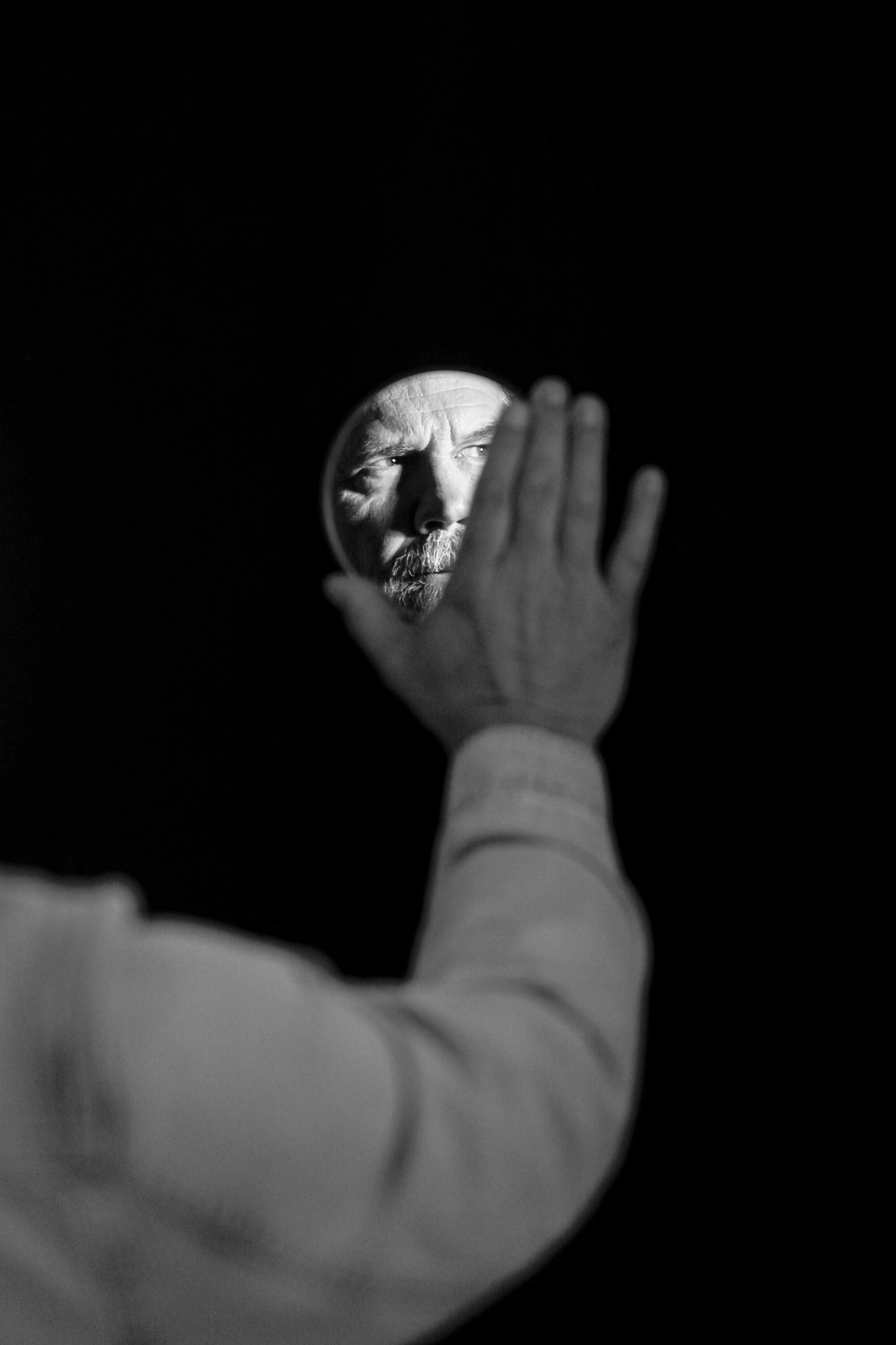 A man holds a mirror against a black background with his face seen in the mirror.