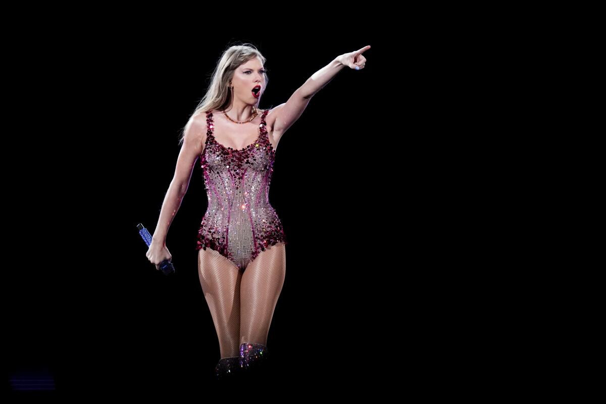 Taylor Swift wears a sparkling red and white leotard and points with her left hand as she performs onstage