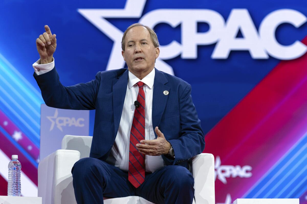 A man sits i a whites chair and points in front of a bright wall that says "CPAC."