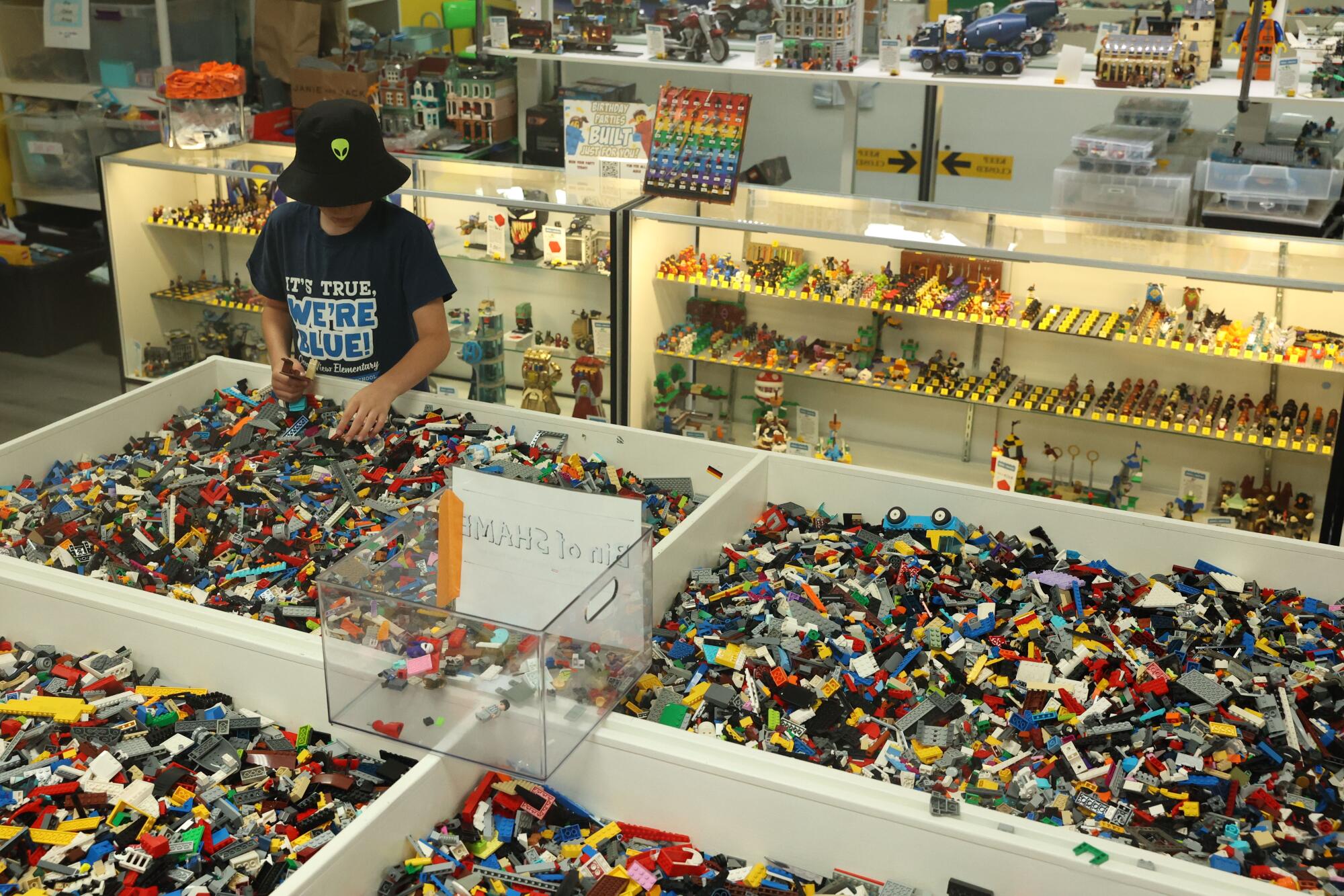 A child wearing a bucket hat browses through bins of Lego pieces near a glass display case
