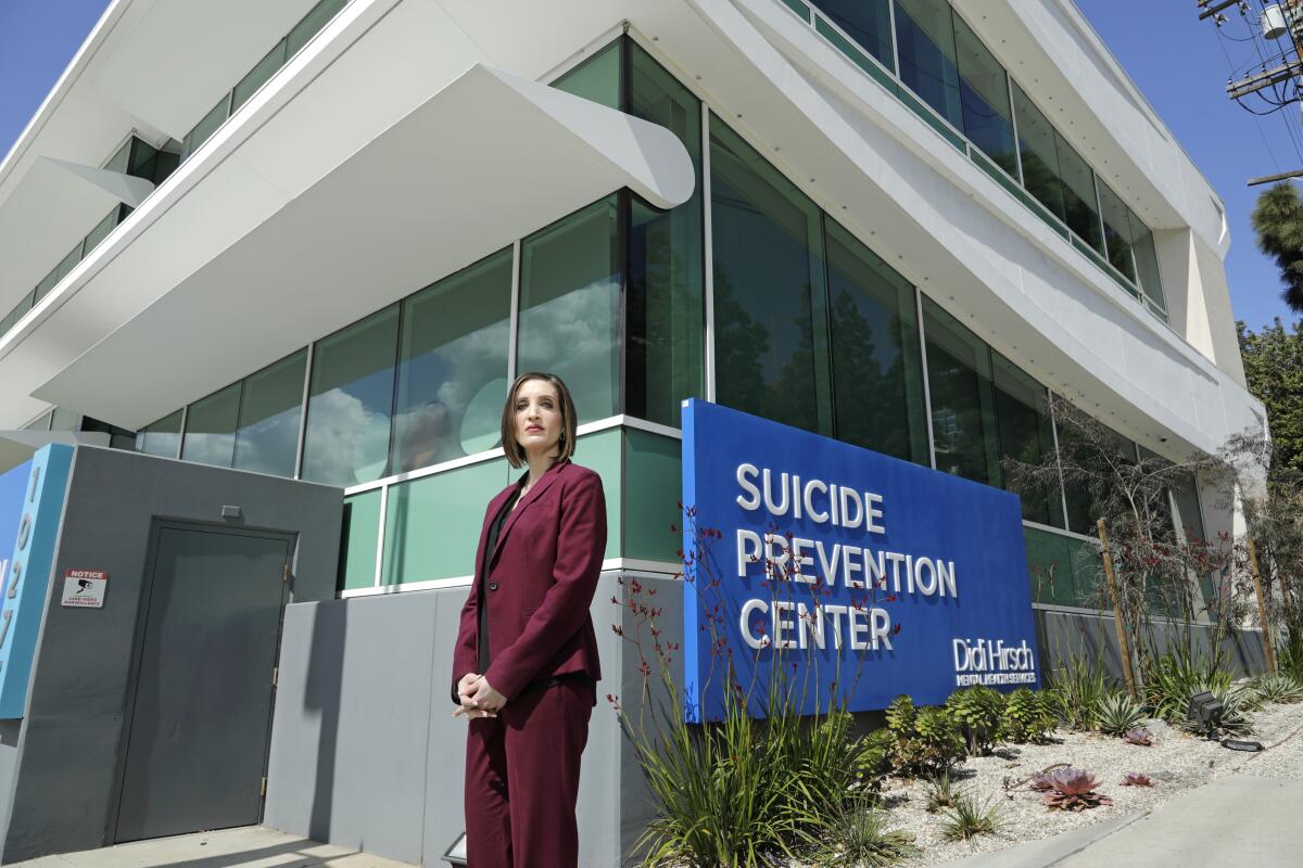 A woman with short hair in a dark red pantsuit stands near a sign that says Suicide Prevention Center outside a building