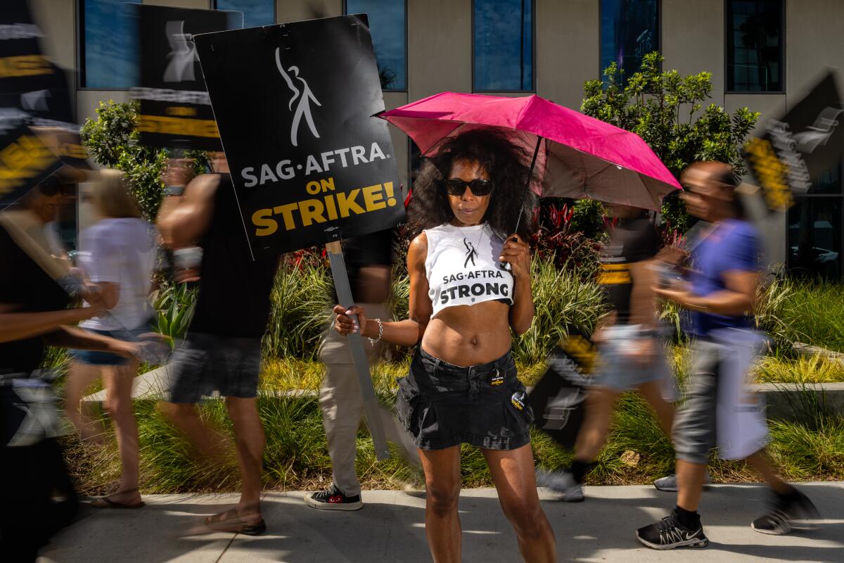 A woman wearing sunglasses and a cropped top holds a SAG-AFTRA sign.