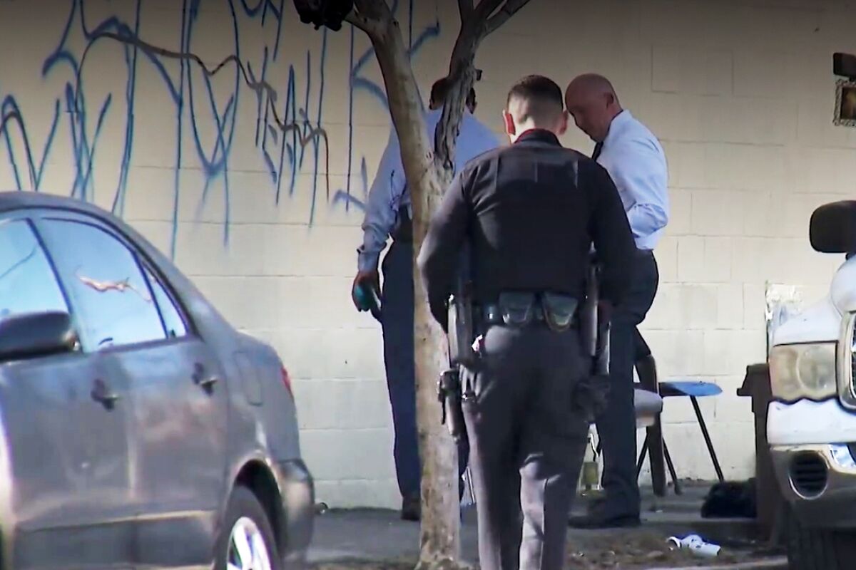 Three people, one in police uniform, stand on a sidewalk near a wall with illegible blue spray-paint writing.