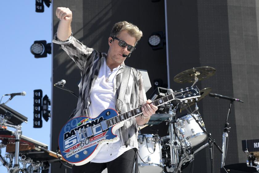 Guitarist Joe Don Rooney in a green button-down and a white shirt holding a guitar on a stage. He is also wearing sunglasses