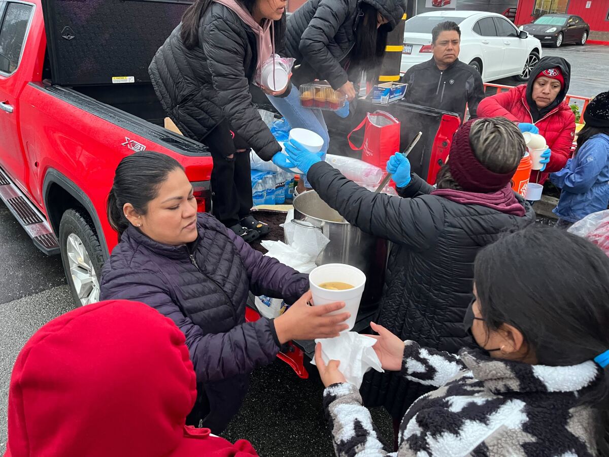 A woman hands  soup to a displaced person as others grab  water from a pickup