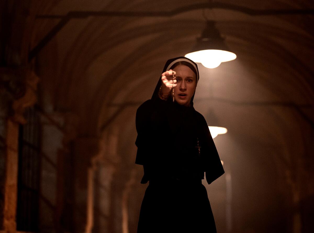 Taissa Farmiga wearing a nun's habit and holding a cross in a dim passageway