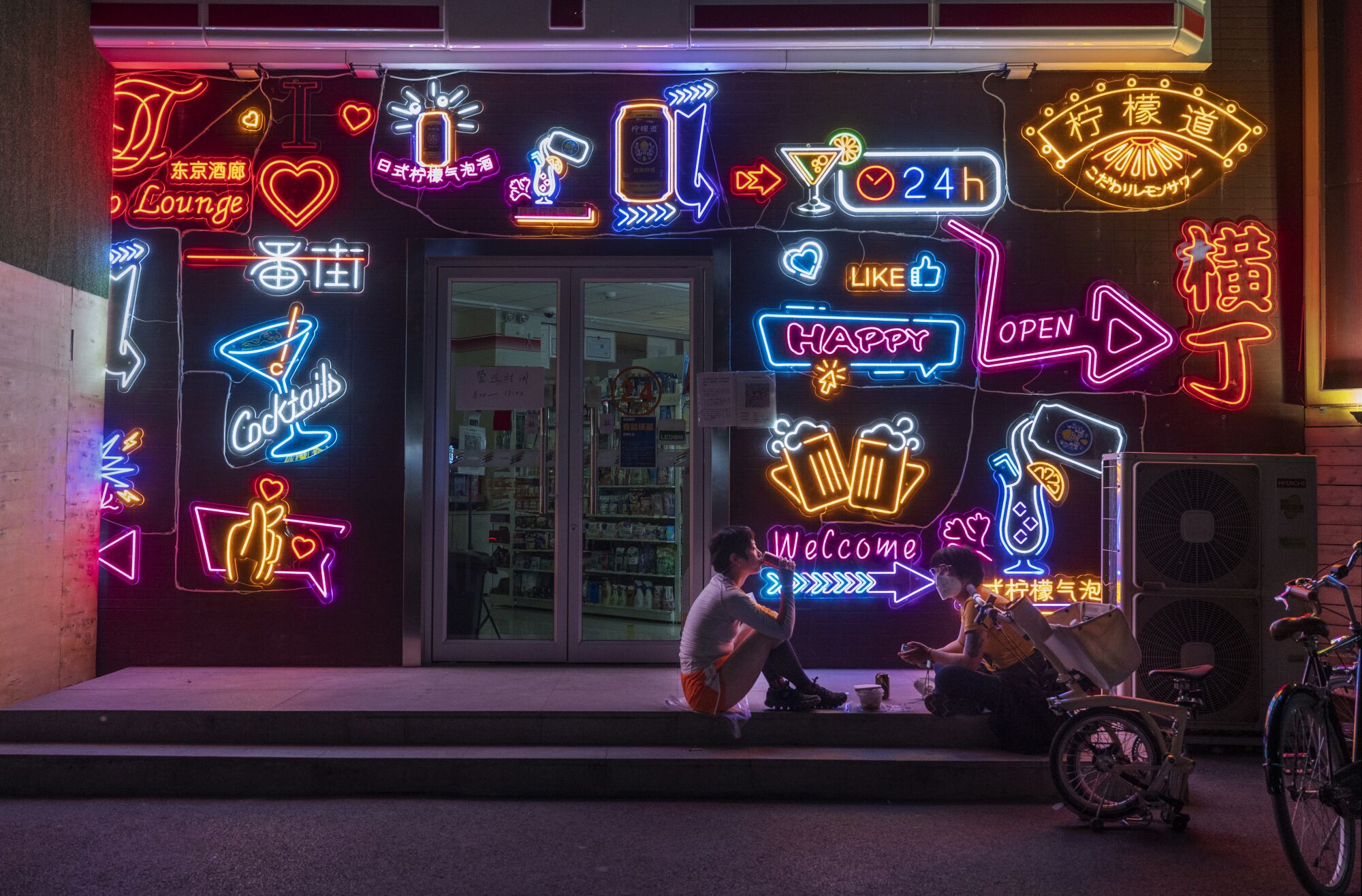 Two people talk on a sidewalk under neon signs.