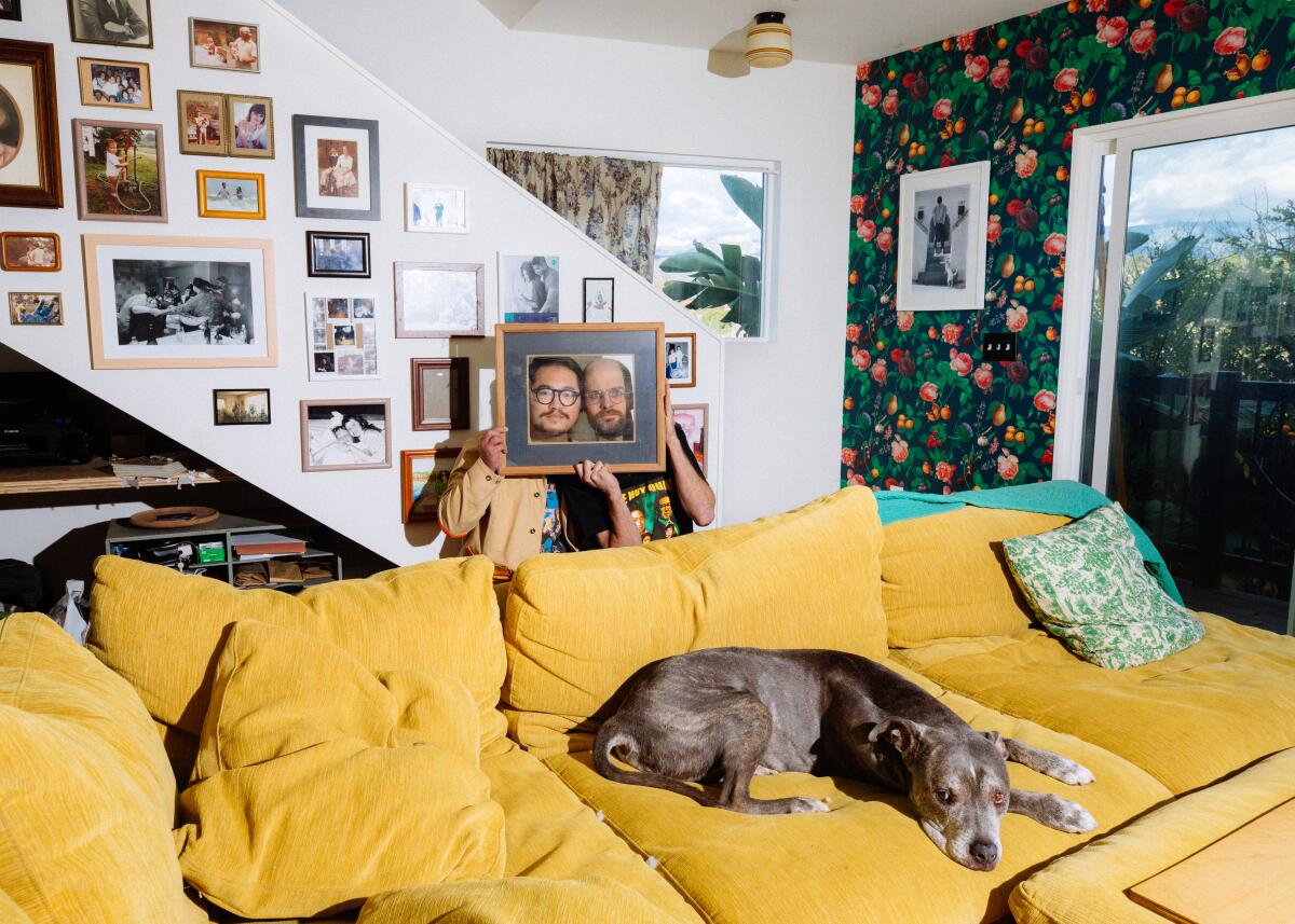 Two men position their faces inside a picture frame as a large dog lounges on the couch.