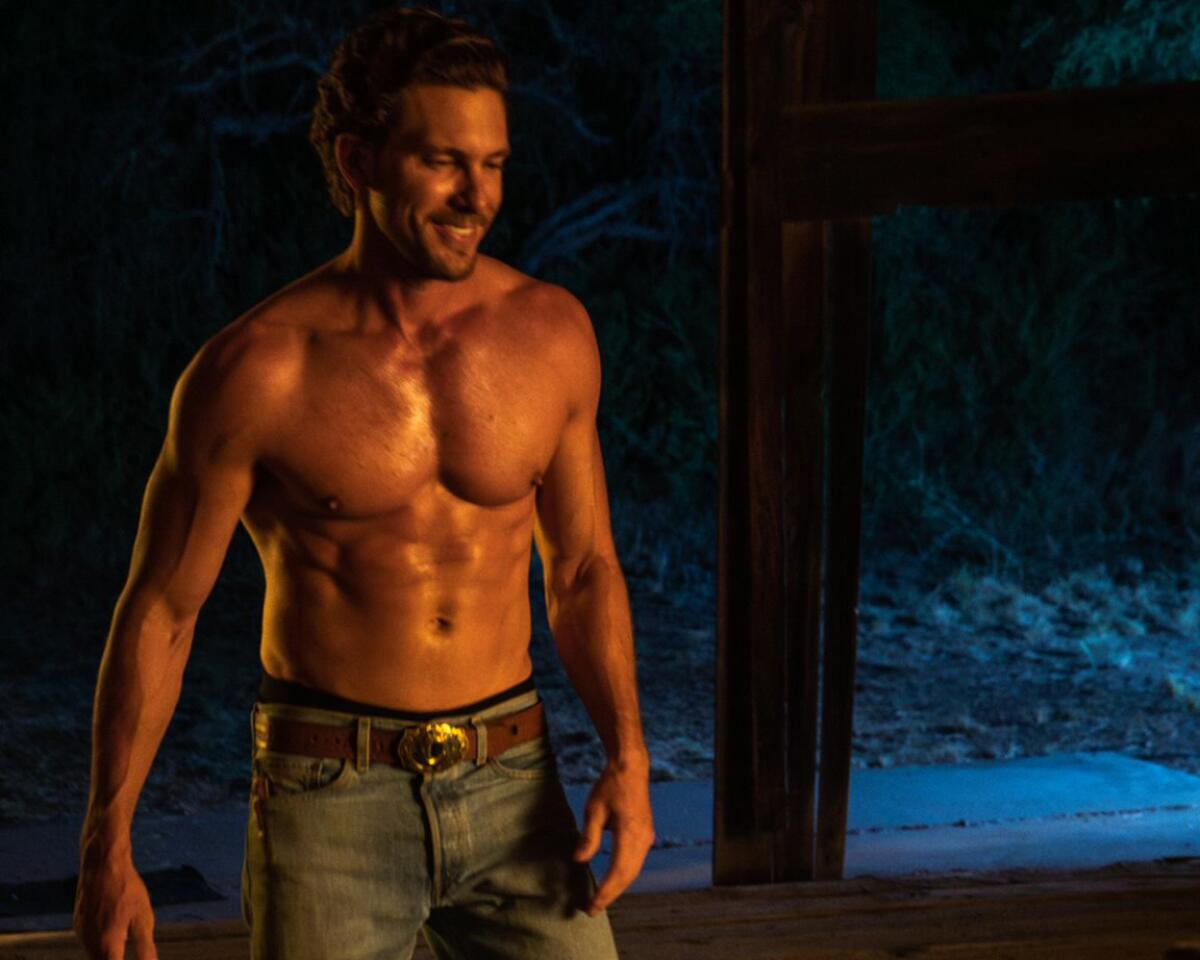 A handsome smiling man wearing no shirt is illuminated by firelight.