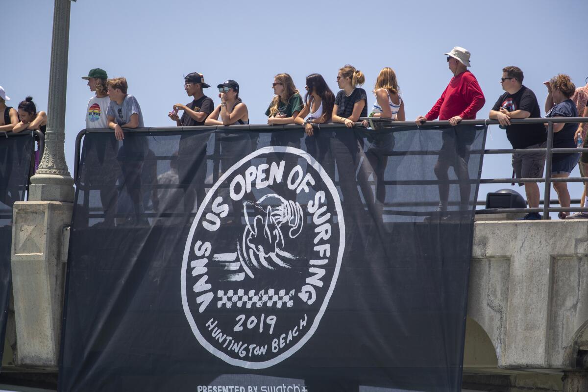 People stand behind a railing where a flag is hung that says "Vans U.S. Open of Surfing."